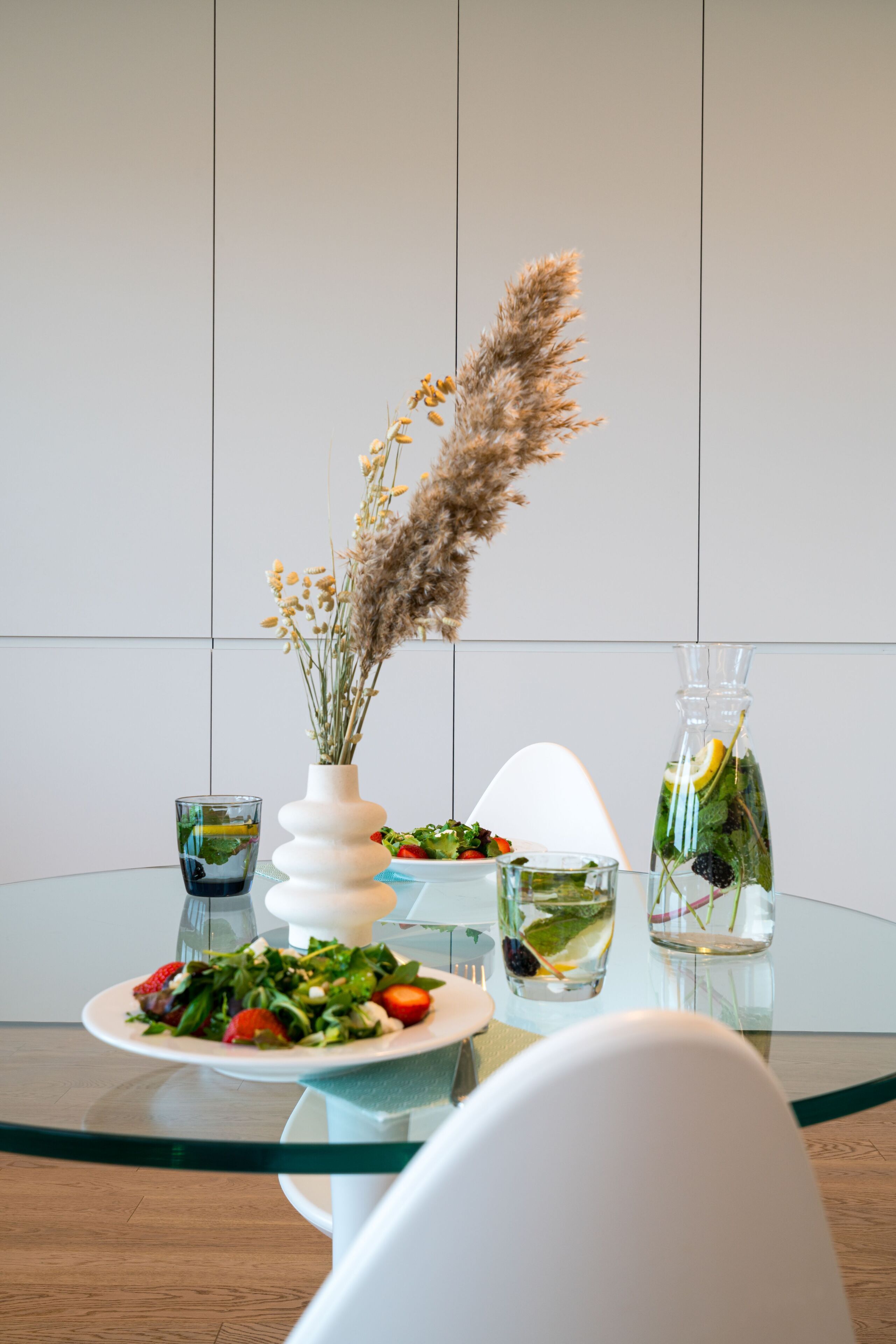 Una mesa de comedor con un plato de ensalada, un vaso de agua, un jarrón con flores secas y una botella de agua con una rodaja de limón.