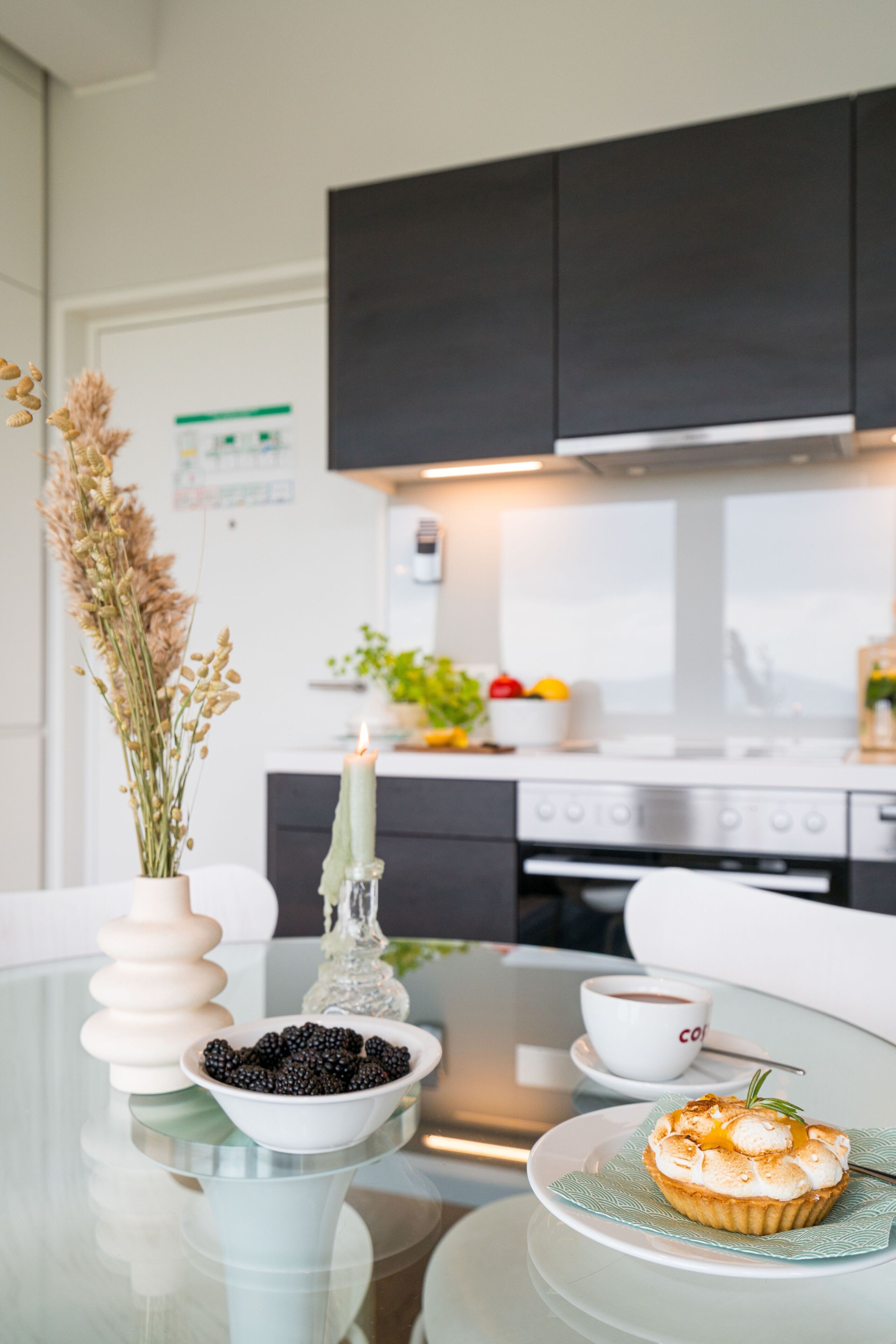 A modern kitchen with dark cabinets, a glass table, a white bowl of blackberries, a lit candle, and a muffin on a plate.