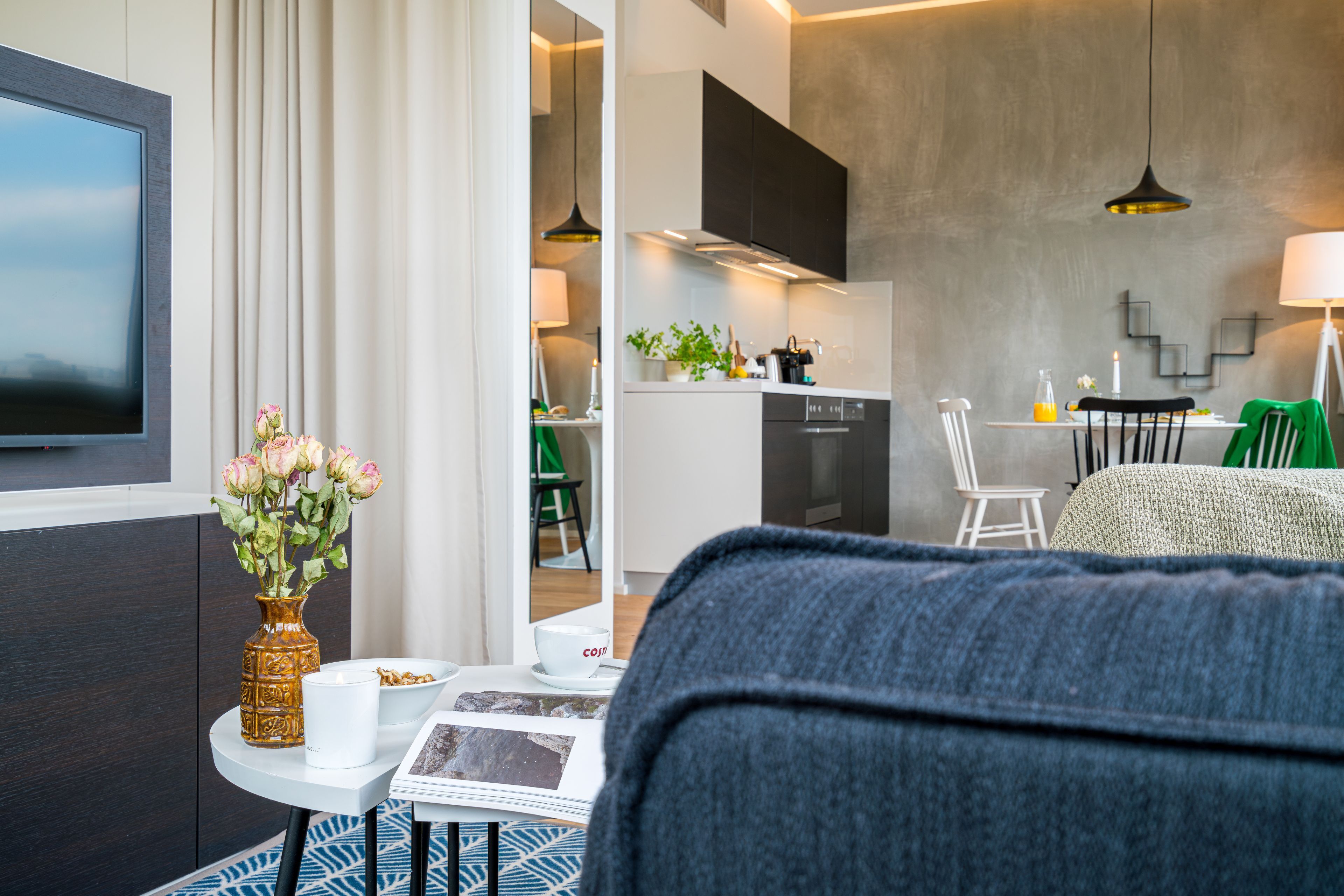 A modern hotel room with a sofa, a small table with a vase of flowers, a flat-screen TV, and a view into the kitchen area.