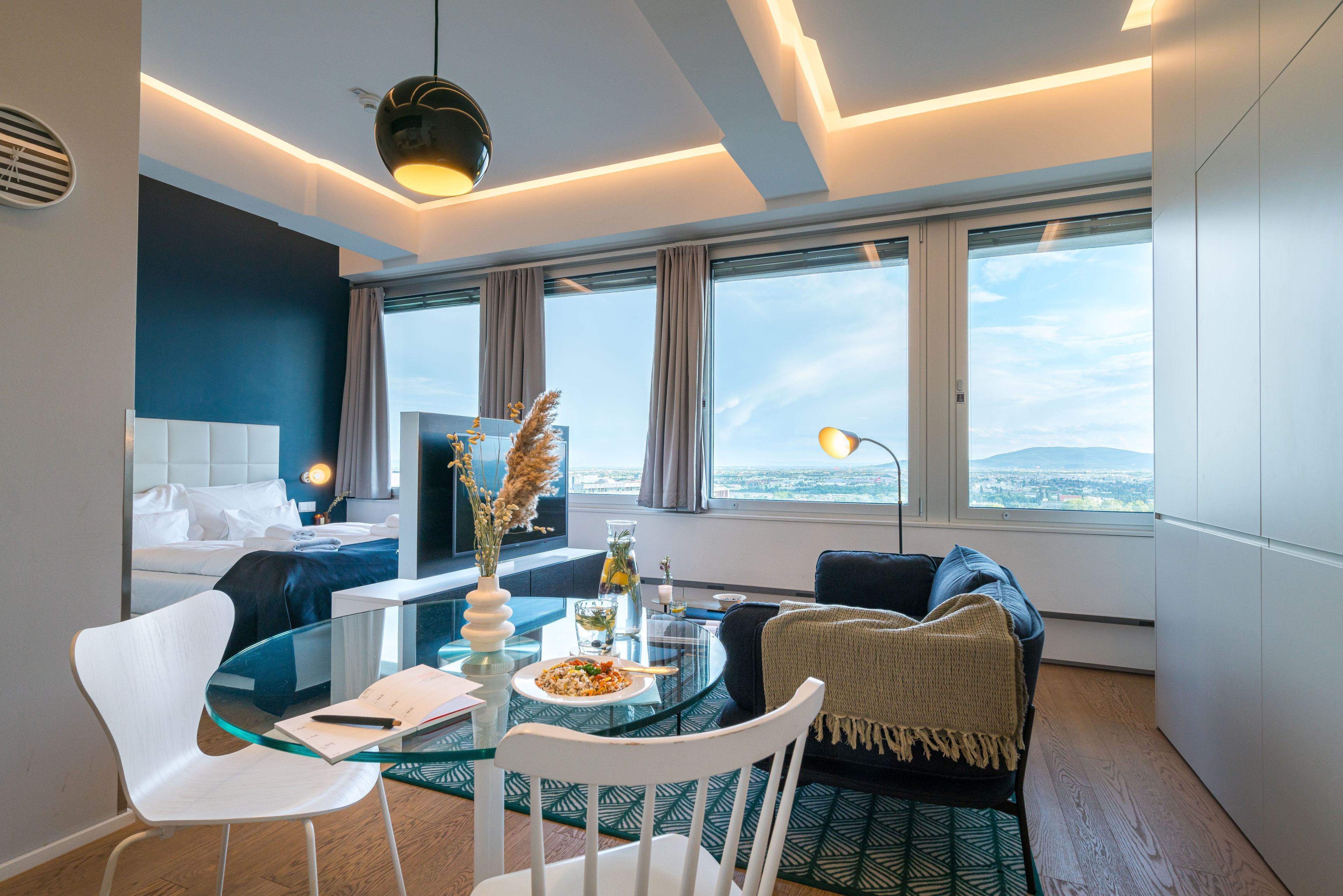 A modern hotel room with a glass dining table, white chairs, a sofa, and a large window offering a city view.