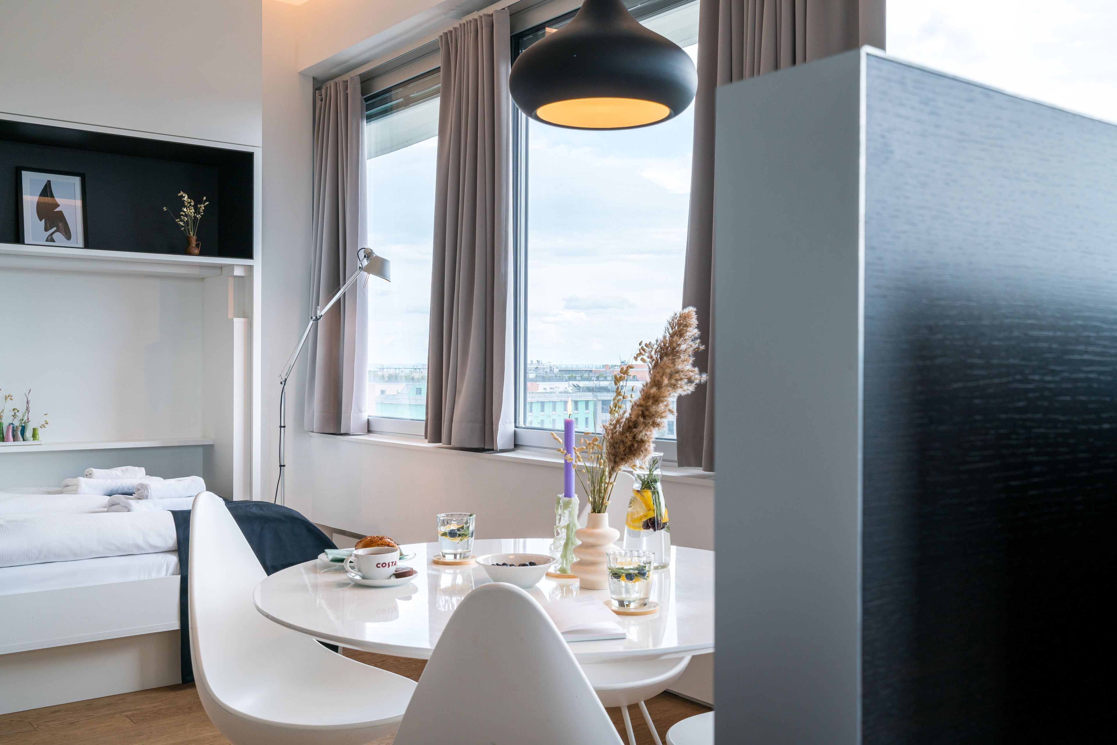 Ein modernes Hotelzimmer mit Bett, Esstisch mit Stühlen, Schreibtisch und großem Fenster mit Vorhängen.
