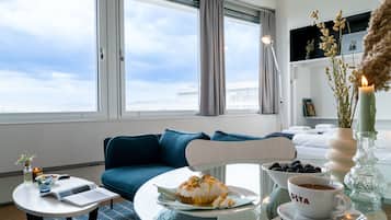 A modern hotel room with a large window, a blue sofa, a dining table with a plate of food, a cup of coffee, and a vase with dried plants.