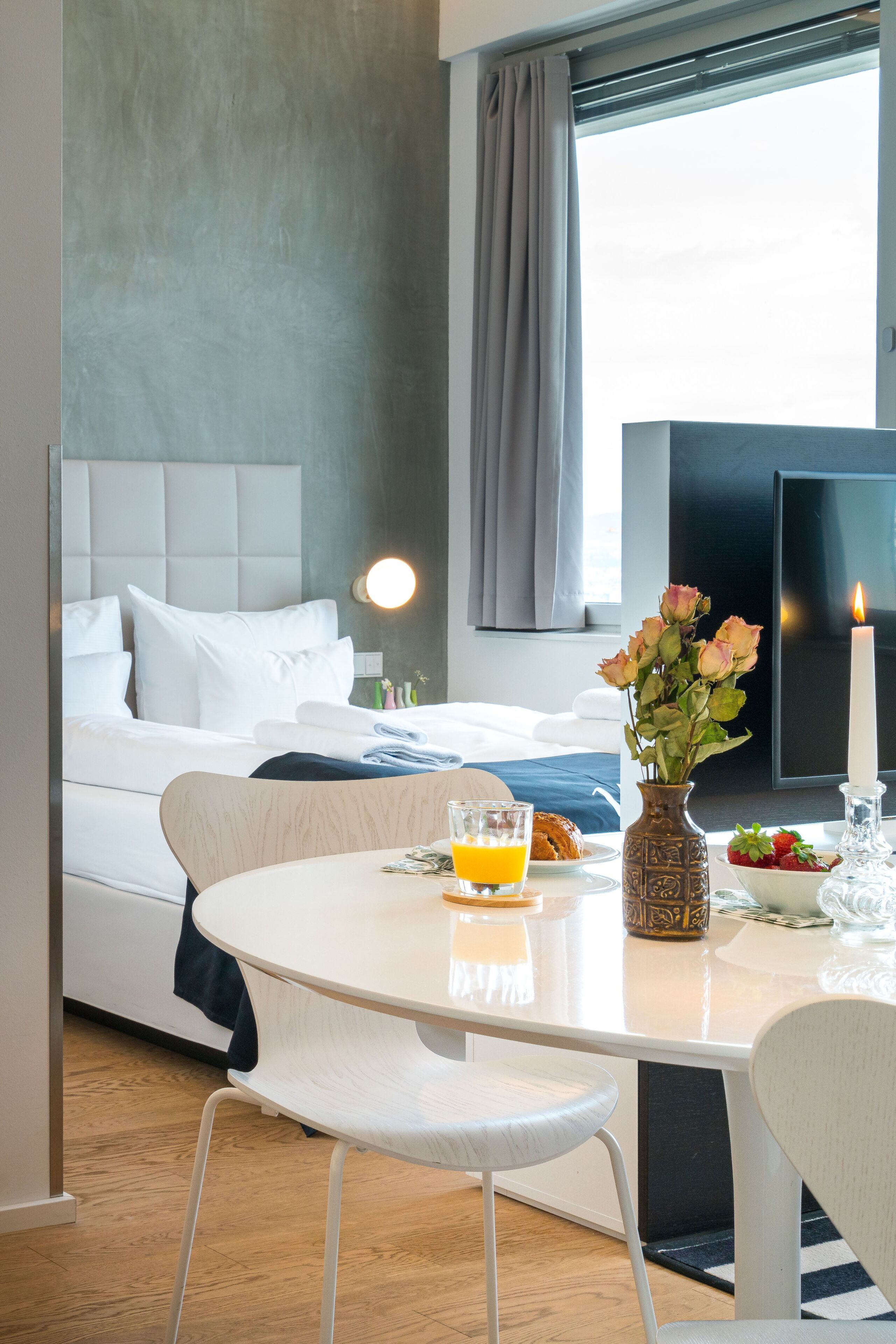 A hotel room with a bed, a bedside table with a vase of flowers, a candle, and breakfast items.
