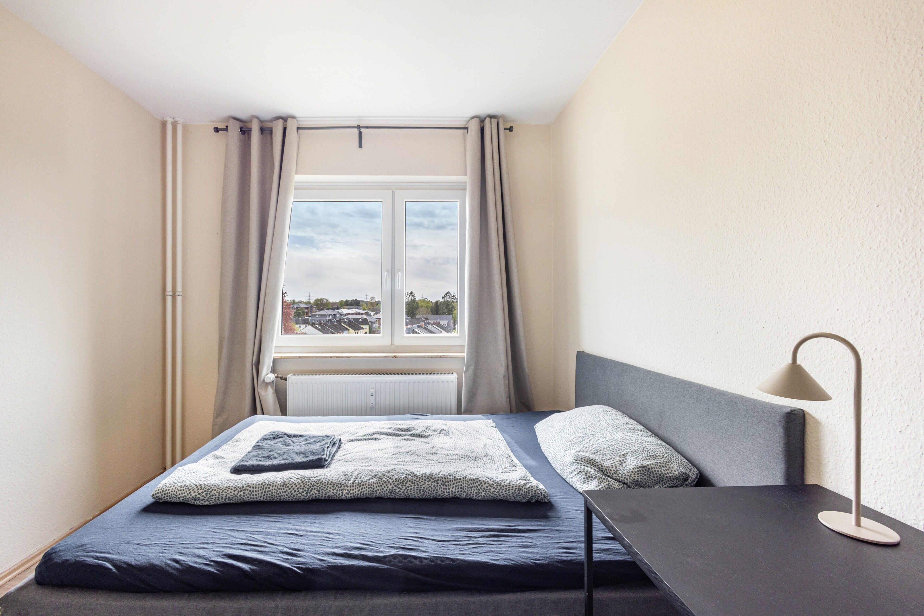 Un dormitorio con cama, mesita de noche, lámpara y ventana con cortinas.