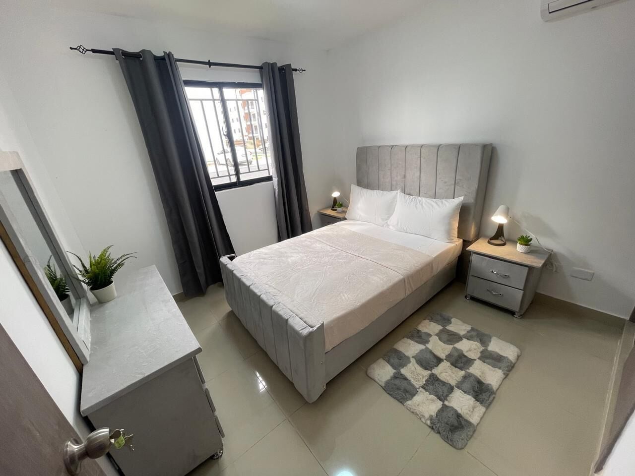 Une chambre à coucher comprenant un lit, des tables de chevet, une commode, un tapis, une plante en pot et une fenêtre avec des rideaux.