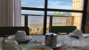 Une table à manger avec des aliments pour le petit-déjeuner, une vue sur un plan d’eau et un bâtiment à la façade quadrillée.