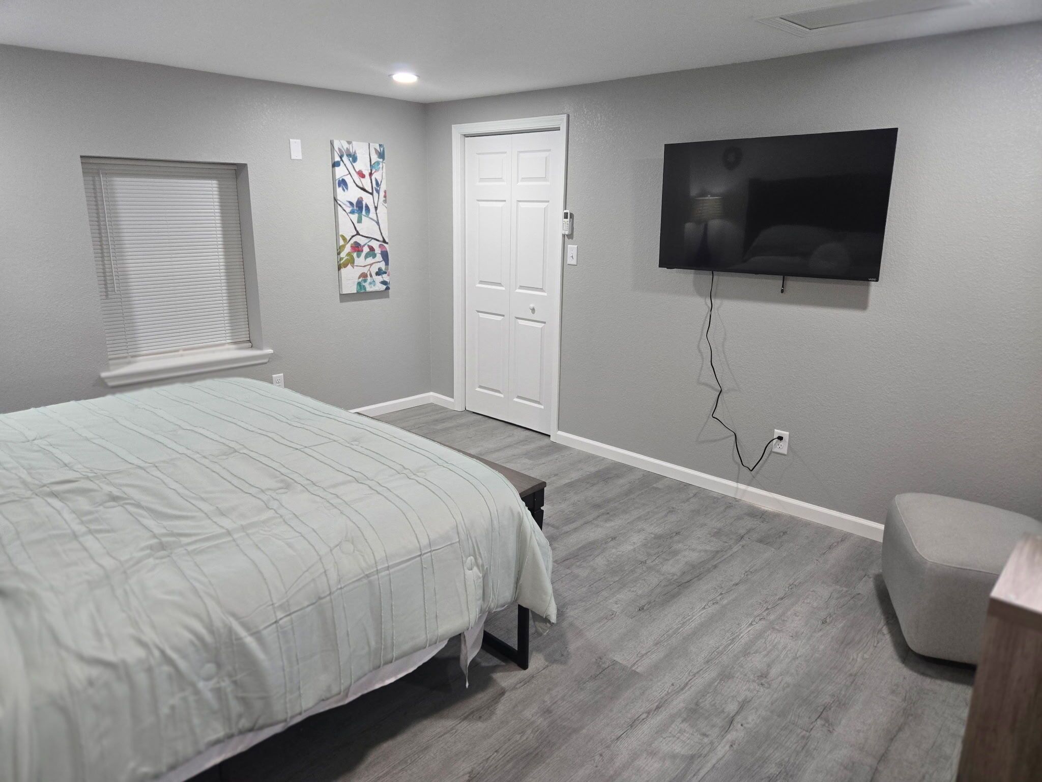 A bedroom with a bed, a bench, a TV, a door, and a window with blinds.