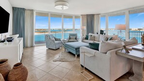 A spacious living room with a large window overlooking a beach, a flat-screen TV, and a comfortable seating area. - Caribe The Resort #C702 | Waterfront, Luxe Resort Amenities, Across from Beach (Orange Beach)