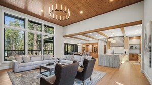 Amplia sala de estar con un sofá grande, dos sillones y una mesa de centro. Cuenta con una cocina con armarios de madera y una isla de mármol. Ventanas amplias ofrecen luz natural y vistas al exterior.