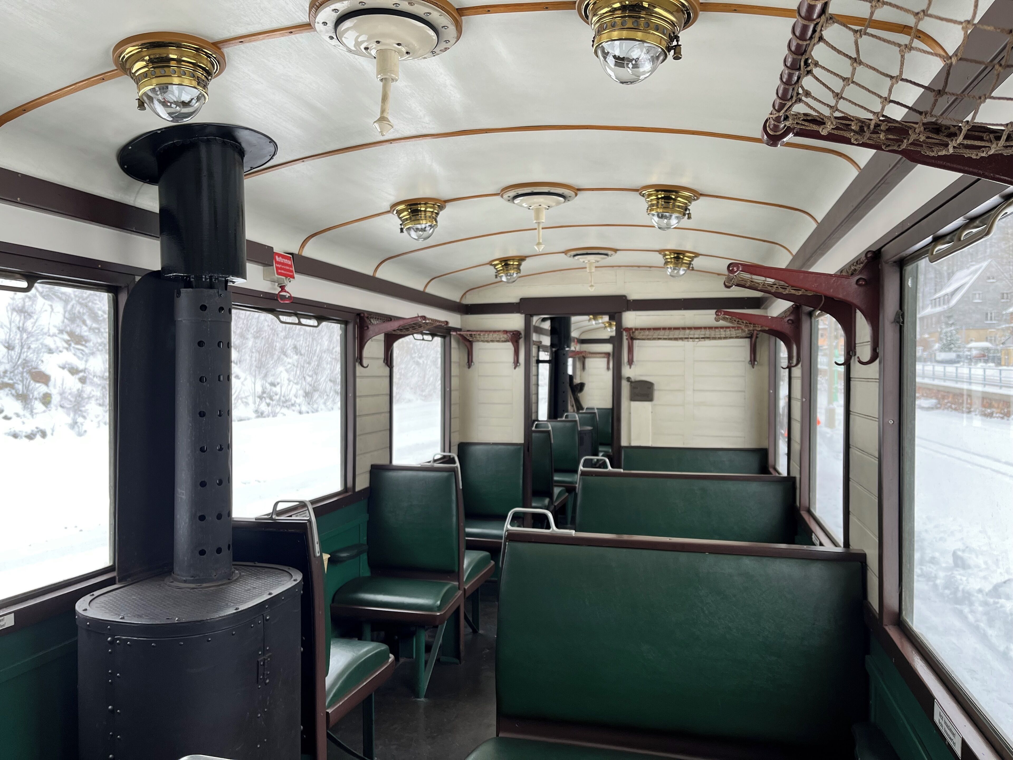 A vintage train interior with green upholstered seats, brass fixtures, and large windows offering a snowy landscape view.