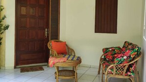 A room with a tiled floor, a wooden door, two wicker chairs, and a small table with a cushion. - Les Ziottines (Deshaies)