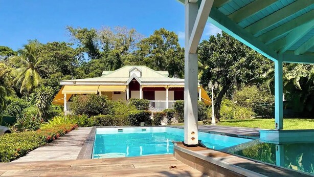 A pool area with a wooden deck, a covered seating area, and a house in the background. - Les Ziottines (Deshaies)