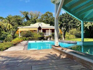 A pool area with a wooden deck, a covered seating area, and a house in the background. - Les Ziottines (Deshaies)
