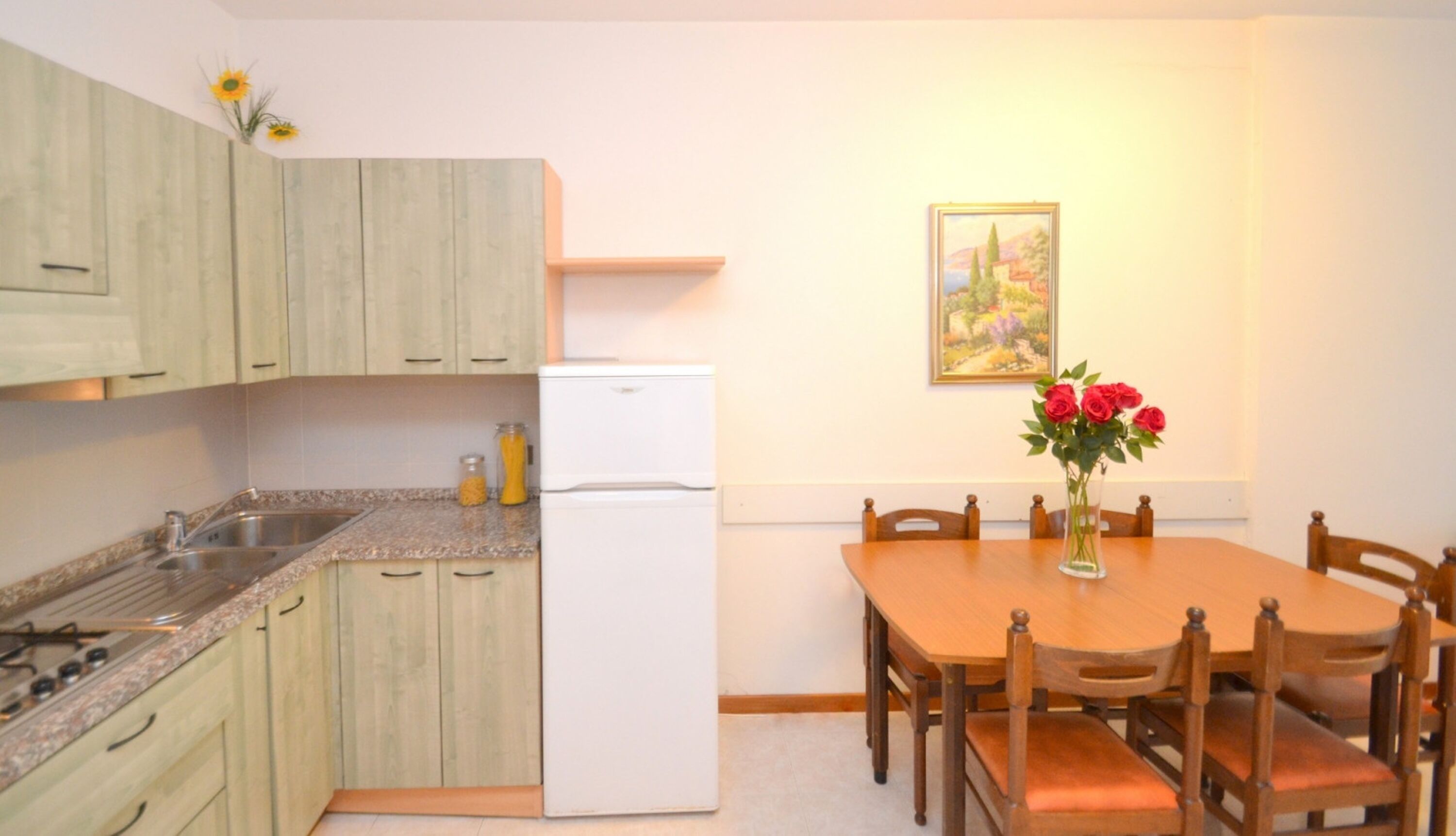 Cocina compacta con armarios de madera, horno integrado y refrigerador. Hay una mesa de comedor con sillas y un florero con flores.
