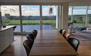 Un espace repas moderne avec une longue table en bois et des chaises noires, de grandes fenêtres offrant une vue sur un paysage verdoyant.