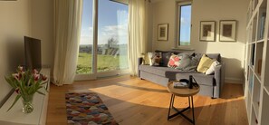 A living room with a sofa, a coffee table, a TV, a bookshelf, and a view of the outdoors. - Stylish home on the Wild Atlantic Way - comfort with space, peace & panorama (Ardfield Clonakilty)