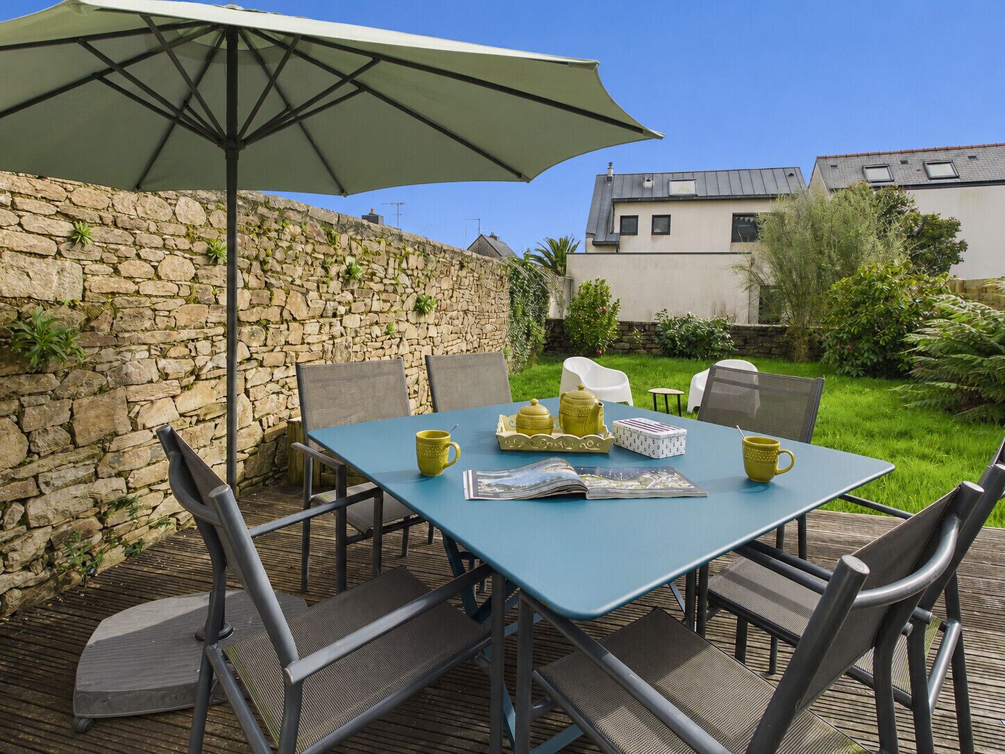 An outdoor patio with a table set for two, chairs, a large umbrella, and a stone wall.