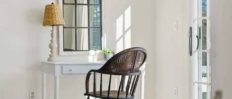 Een wit bureau met een geweven lamp, een donker houten stoel en een spiegel met een decoratieve lijst.