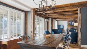 A rustic dining area with a wooden table and chairs, a chandelier, and a view of a living room with blue chairs and a television. - Spacious Farmhouse Near the Beach Circa 1860 (Biddeford)