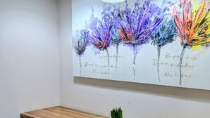Un área de comedor con una mesa de madera, dos sillas negras, una pequeña planta en maceta y un cuadro abstracto colorido en la pared.
