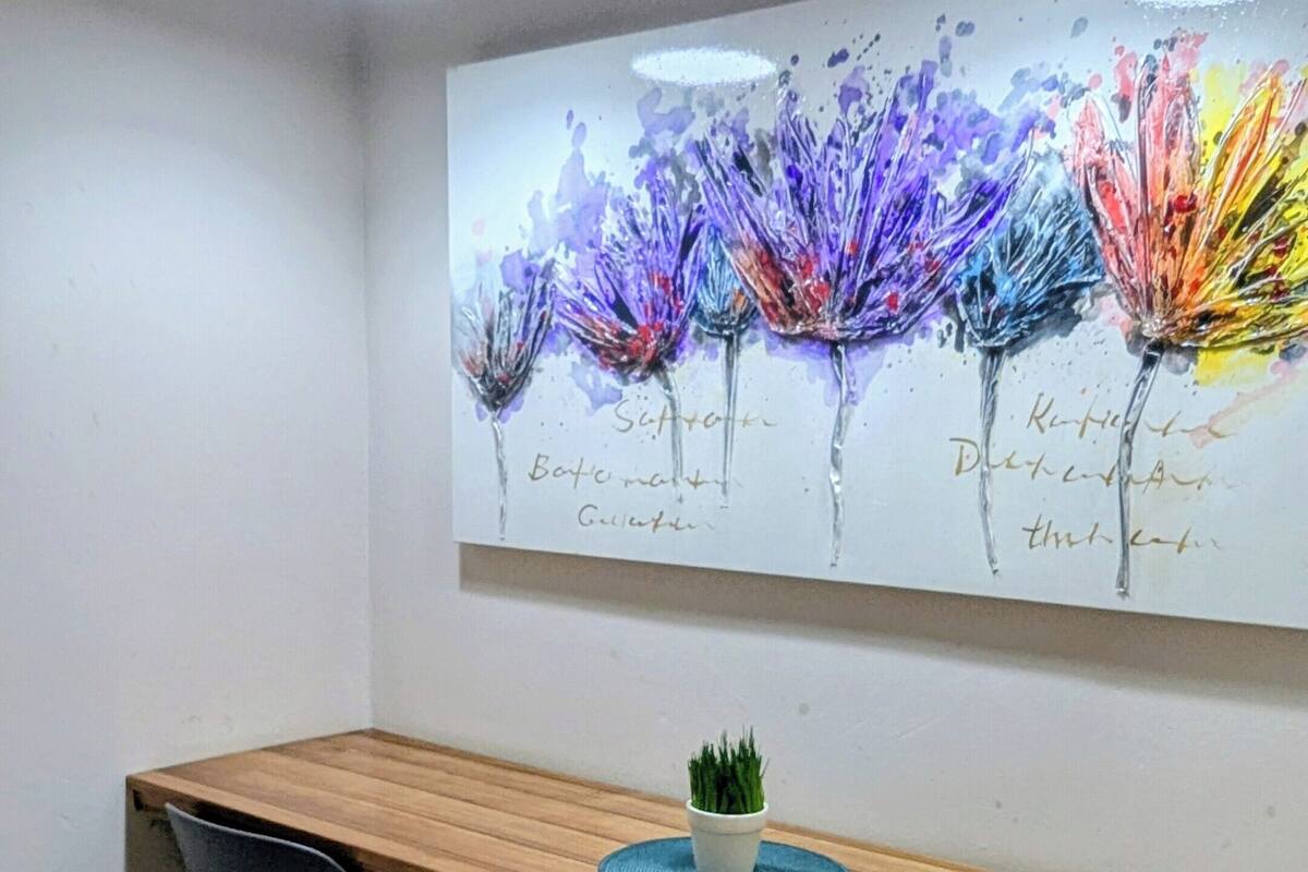 Un coin repas avec une table en bois, deux chaises noires, une petite plante en pot et un tableau abstrait aux couleurs vives accroché au mur.