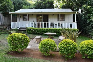Uma casa térrea com varanda, cercada por sebes bem cuidadas e um pequeno jardim com mesa e cadeiras de pedra.