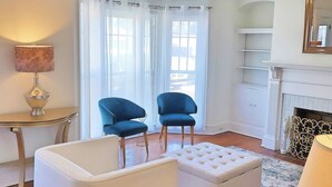 A modern living room with a chandelier, two blue armchairs, a white ottoman, a small table, and a fireplace. - Charming Home on Porter Avenue! (Biloxi)