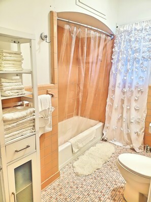 A bathroom with a bathtub, a toilet, a shelf with towels, and a shower curtain. - Charming Home on Porter Avenue! (Biloxi)