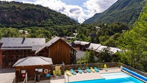 Zona de piscina con un edificio de madera, sillas de descanso y montañas al fondo.