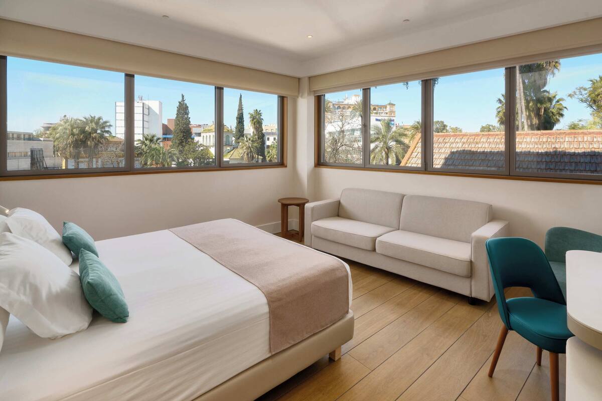 A modern hotel room with a bed, a sofa, a small table, and large windows offering a city view.