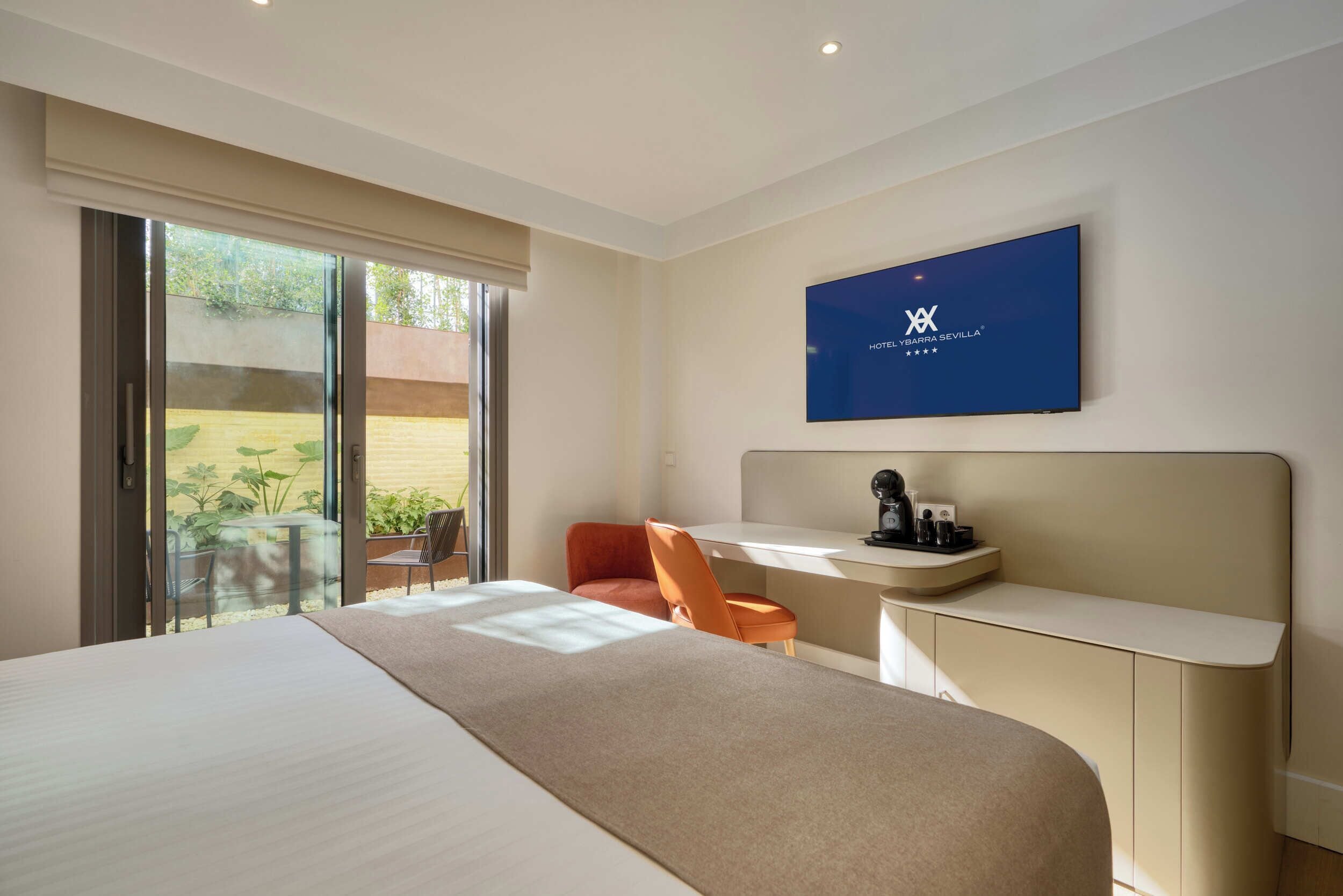 Een moderne hotelkamer met een groot bed, een bureau met stoel, een televisie en een glazen schuifdeur naar een buitenruimte.