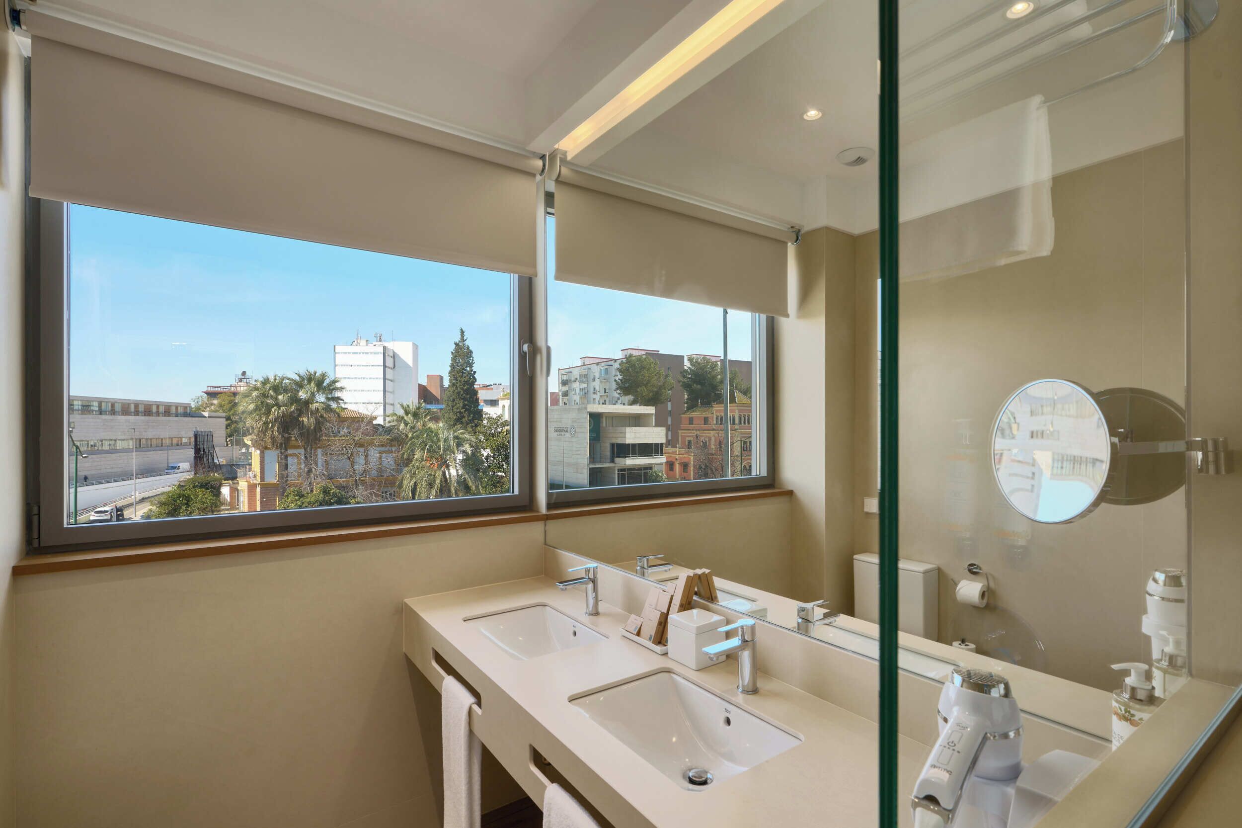 A modern bathroom with dual sinks, a large mirror, and a view of the cityscape through a large window.