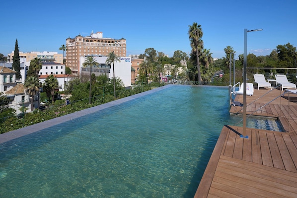 A clear pool with a wooden deck, lounge chairs, and a hospital building in the background. - Hotel Ybarra Sevilla (Seville)