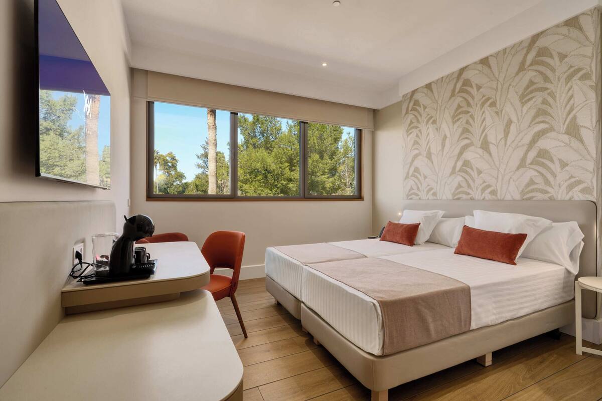 A modern hotel room with a large bed, a desk with a chair, and a window with a view of trees.