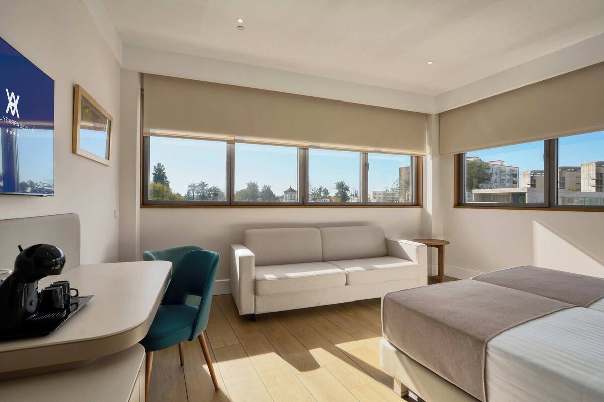 A modern hotel room with a sofa, a bed, a dining area with a table and chairs, and large windows offering a city view.