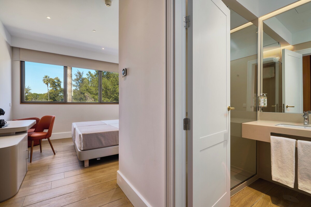 A modern hotel room with a bed, a desk, a chair, a bathroom with a mirror, and a view of trees through large windows.