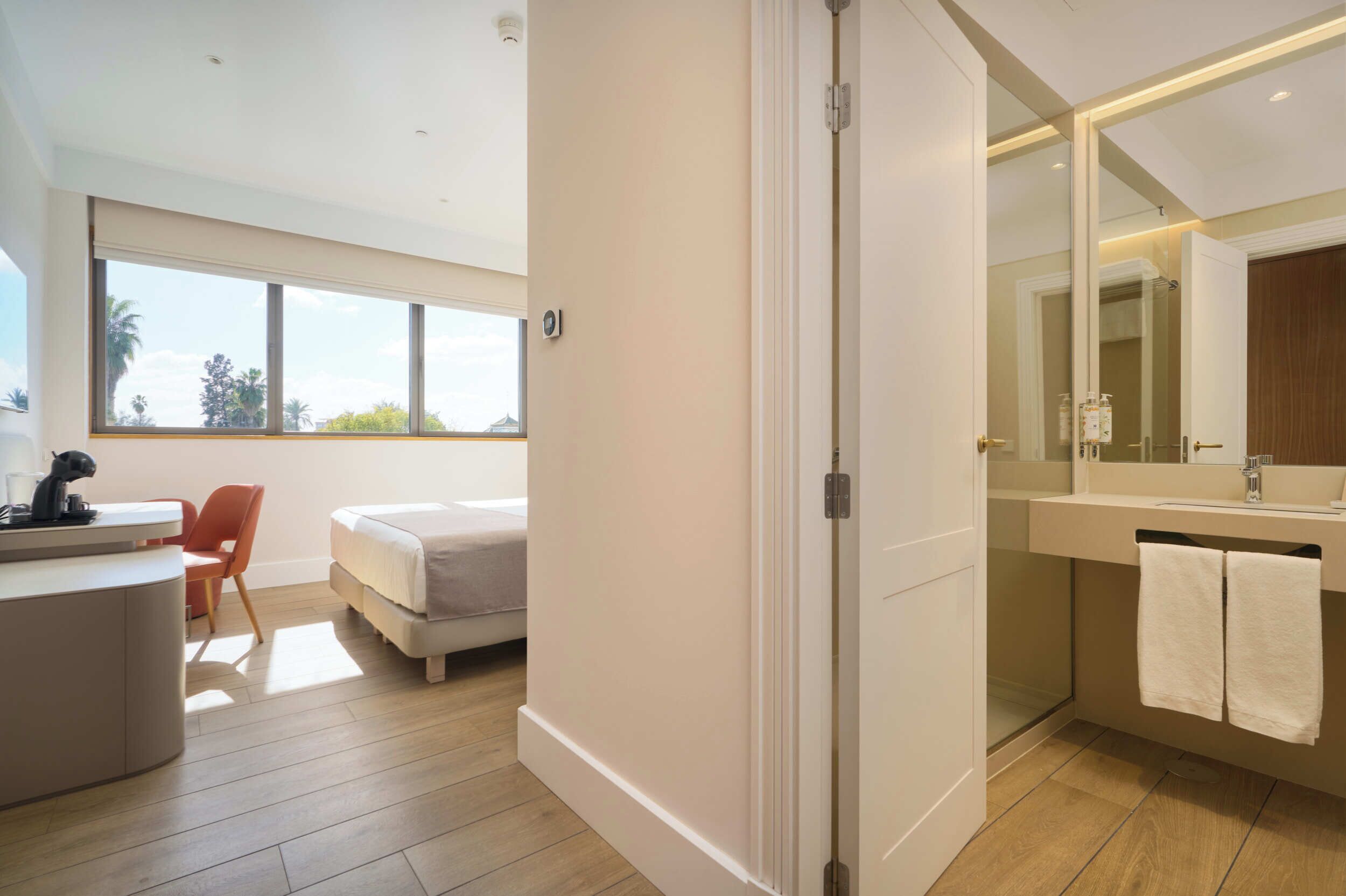 Una habitación de hotel moderna con cama, escritorio, silla y un baño grande con espejo y toallero.