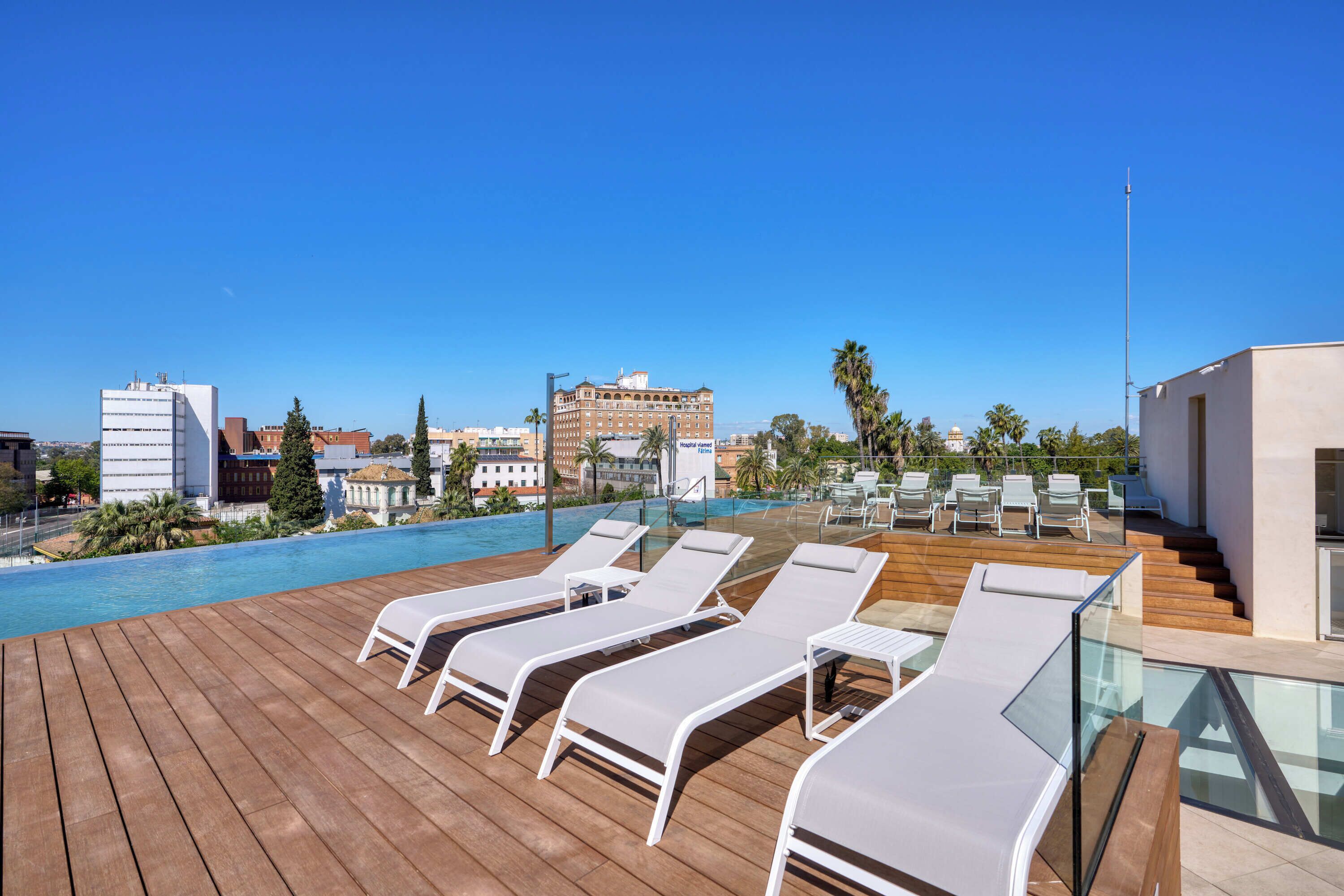 A rooftop pool area with lounge chairs, a clear blue sky, and buildings in the background.