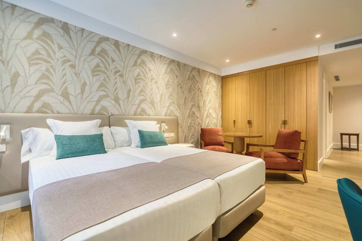 A hotel room with a large bed, two chairs, a small table, and a wooden wardrobe.