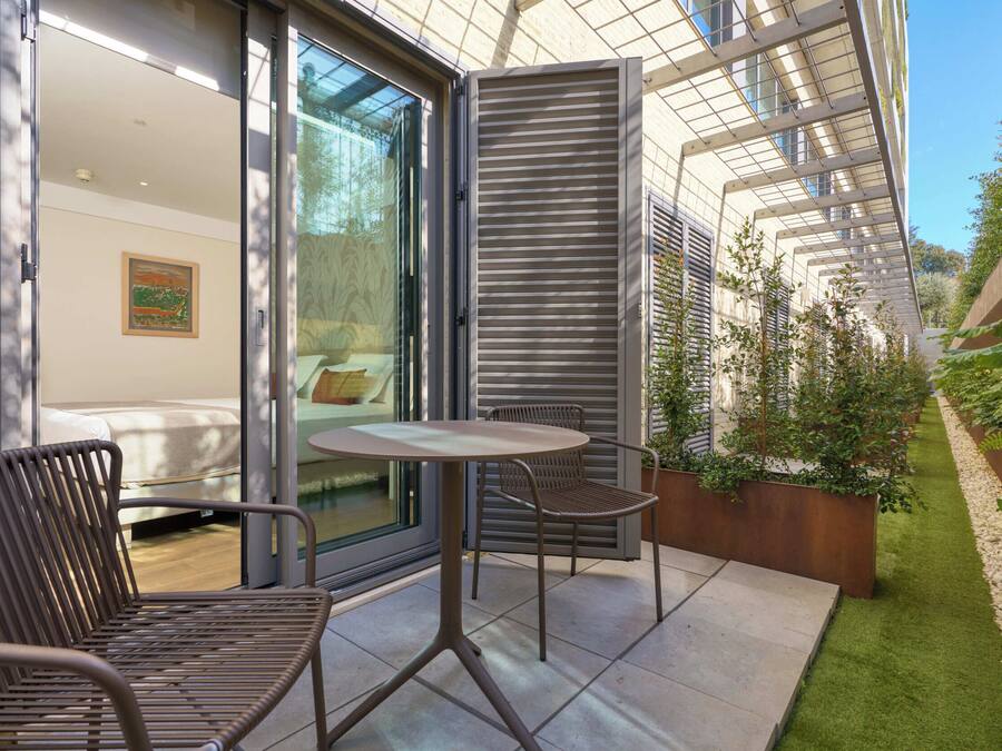 Een modern terras buiten, met een tafel en stoelen, een schuifdeur naar een kamer en een kleine tuin met planten.