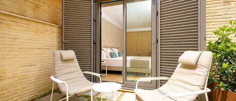 Un espace extérieur moderne avec deux chaises et une petite table, une porte-fenêtre coulissante donnant accès à une chambre, et un mur en briques.