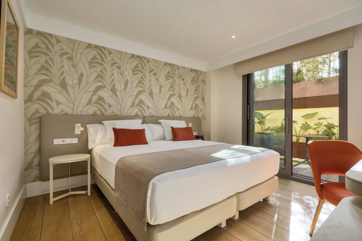 A bedroom with a bed, a small bedside table, a chair, and a large sliding glass door to the outside.