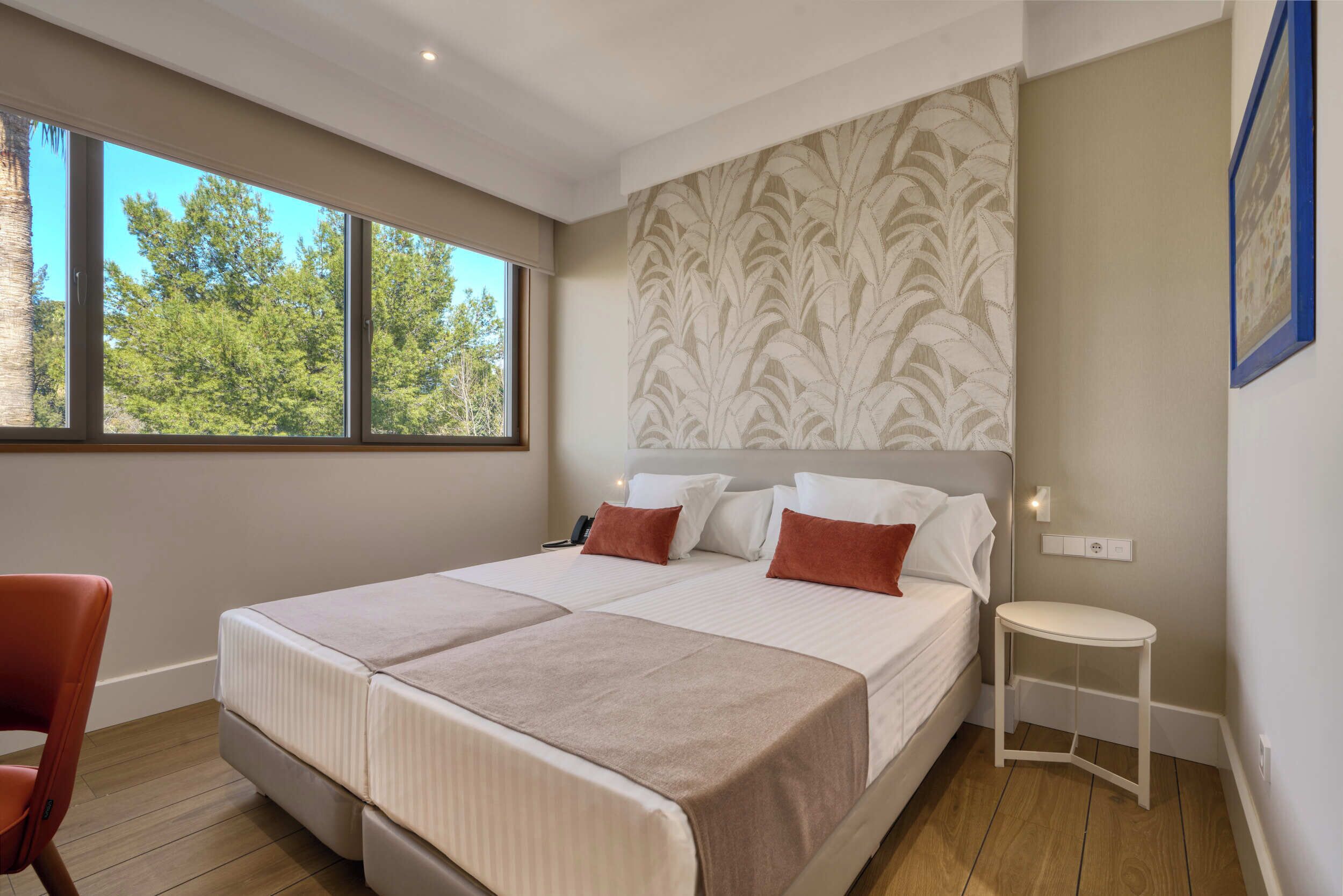 A modern hotel room with a large bed, a small side table, a chair, and a window with a view of trees.
