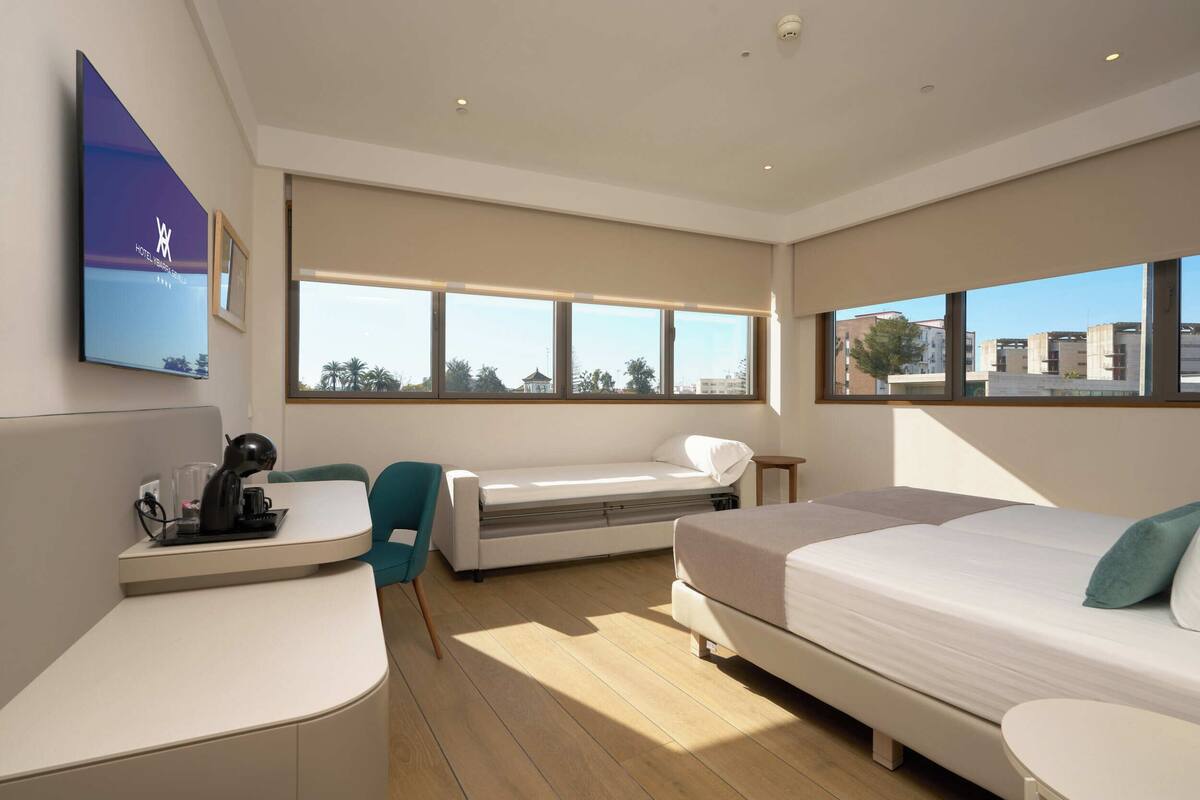 A modern hotel room with a bed, a desk, a chair, and large windows offering a city view.