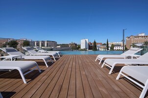 Terraza de madera con sillones blancos frente a una piscina, rodeada de edificios.