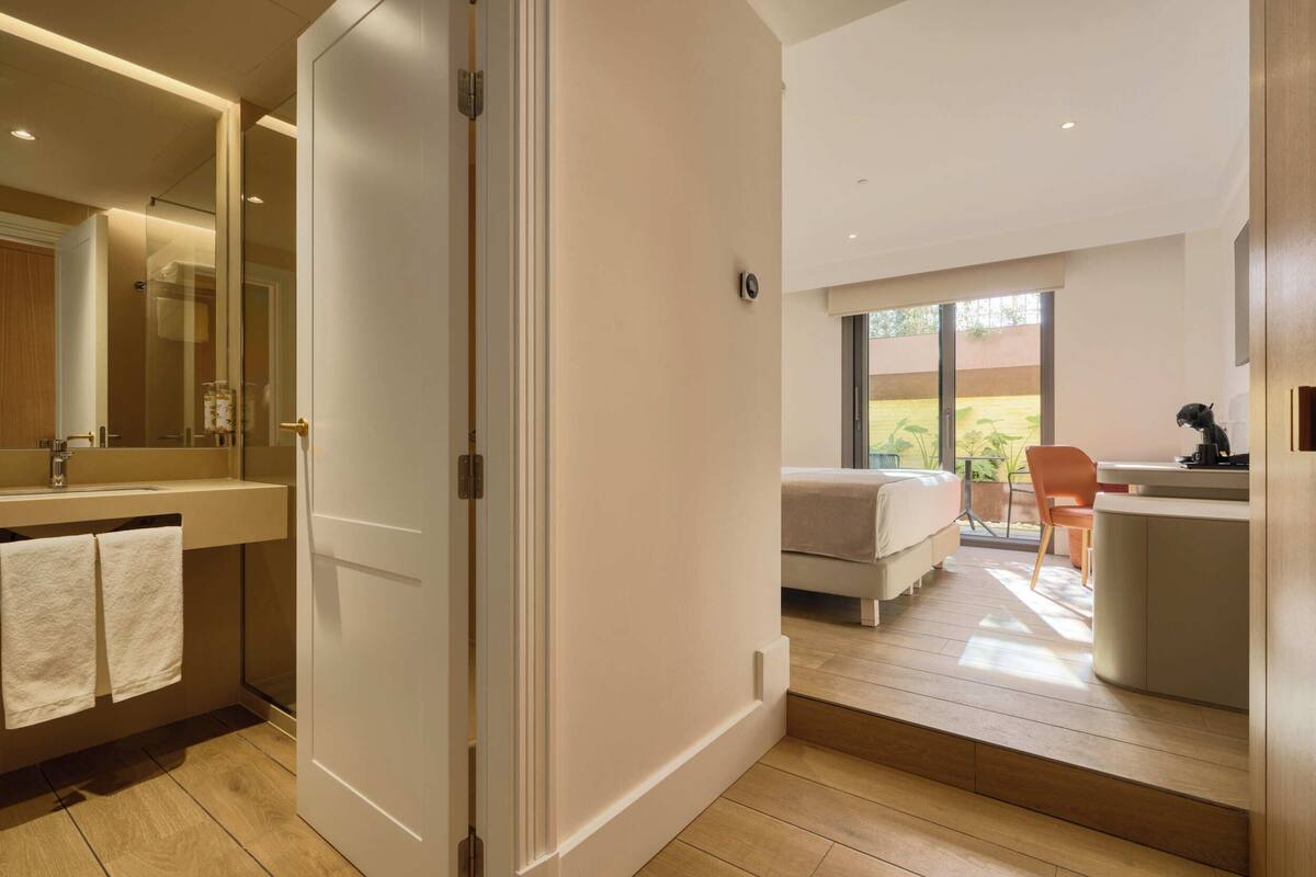 A modern hotel room with a large mirror, a bed, a desk, and a view of a garden.