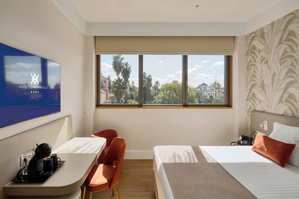 A hotel room with a bed, a desk, a chair, a TV, and a window with a view of palm trees and buildings.