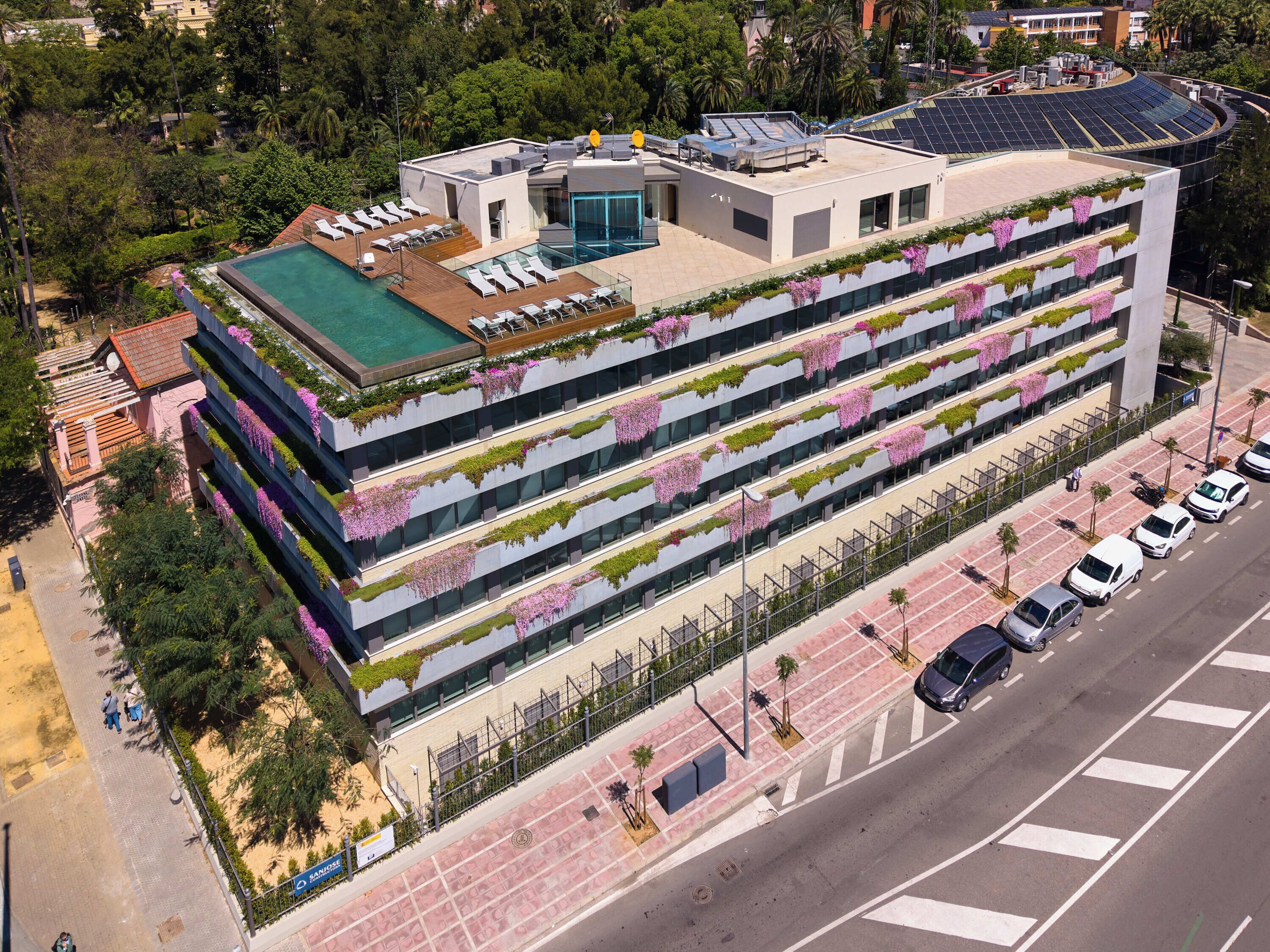 Un immeuble à plusieurs étages doté d’une piscine sur le toit, de balcons ornés de plantes suspendues et offrant une vue sur la rue, avec ses voitures et ses piétons.