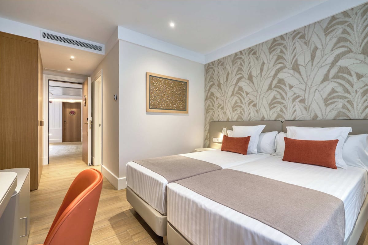 A hotel room with two beds, a desk, a chair, a wall-mounted artwork, and a patterned wallpaper.