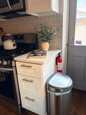 A kitchen with a stove, oven, white cabinets, a potted plant, a book, and a trash can. - River Place -Walk to Jefferson Methodist, 2BR Queen Beds 1 Bath -Patio & Grill (Philadelphia)